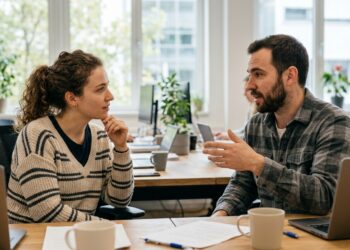 Two colleagues in an office, one leaning in and listening attentively while the other speaks